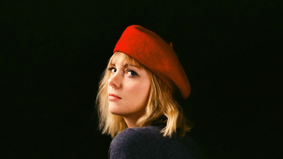 Photo of Saskia Keultjes, a woman with blond hair wearing a red beret looking back over her shoulder against a black backdrop