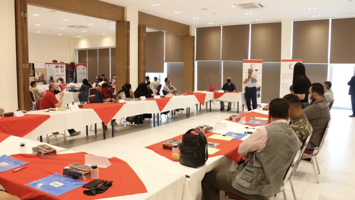 Wide angle photo of a meeting room with people sitting at long tables listening to a lecture