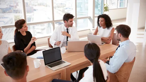 5 people sitting around a conference table with laptops in discussion