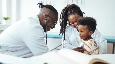 Photo of doctor using stethoscope to listen to little boy's heart who is sitting on a woman's lap