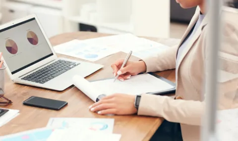 Image of person sitting at a wooden desk with a laptop open and taking notes on a legal pad