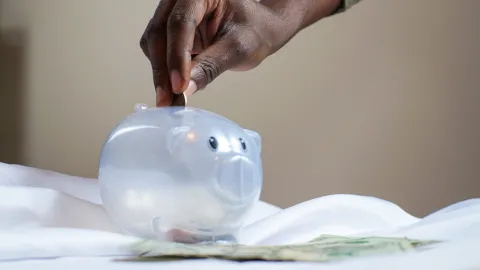 Person's hand putting a coin into a clear plastic piggy bank