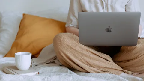 photo of person sitting on top of a bed with a laptop and a coffee cup